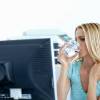 Blondierte Frau in türkiser Bluse trinkt vor einem Bildschirm sitzend aus einem Glas Wasser