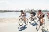 Drei Triathleten und eine Triathletin trainieren auf dem Rennrad auf einer sonningen Strasse am Wasser