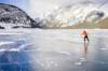 Frau in oranger Jacke fährt Schlittschuh auf zugefrorenen Gebirgssee