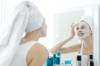 Weisse Frau in weissem Badezimmer mit weissem Handtuchturban steht vor dem Badezimmerspiegel und kontrolliert ihre weisse Gesichtsmaske 