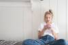 Blondes Teenie-Mädchen mit wiessem T-Shirt und Jeanshose sitzt auf dem Bett, an die weisse Holzwand gelehnt und hält mit beiden Händen sein Smartphone, in das es konzentriert blickt