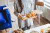 Frau in grauem Pullover kommt mit einem Brettchen auf dem ein Orangensaft, Muesli und ein Croissant steht zum Frühstückstisch