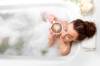 Von oben gesehen: Frau liegt entspannt in schaumiger Badewanne und hält sich einen Cappuccino mit Milchschaumherz an die Wange