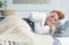 Rothaarige Frau liegt mit Häkeldecke und Bademantel im Bett und schaut besorgt auf ihr Fieberthermometer