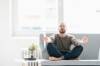 Mann mit rundem Gesicht, dünnem Vollbart, geschlossenen Augen und blossen Füssen sitzt im Schneidersitz auf einem Sofa, die Arme ausgestreckt - neben sich auf einem Tischchen ein Bonsai - und tut so, als wenn er meditiert