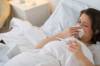 Frau liegt im Bett und schaut beim Naseputzen auf ihr Fieberthermometer