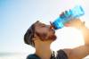 Junger Mann mit Flaumbart und Mützchen trinkt aus blauer Plastikflasche Wasser, Sonne im Hintergrund
